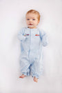 A baby with light hair and blue eyes lies on a white surface, wearing The Beaufort Bonnet Company’s Twinkle Twinkle 2-in-1 - Buckhead Blue with Trains romper in soft Pima cotton, featuring small embroidered train designs on the chest. The baby is looking up and smiling slightly.
