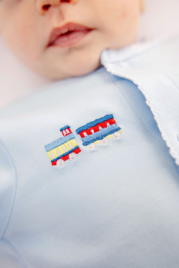 Close-up of a baby wearing The Beaufort Bonnet Company’s Twinkle Twinkle 2-in-1 romper in Buckhead Blue with embroidered yellow and blue trains on the chest. The baby’s mouth and part of the face are visible.