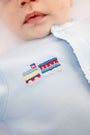 Close-up of a baby wearing The Beaufort Bonnet Company’s Twinkle Twinkle 2-in-1 romper in Buckhead Blue with embroidered yellow and blue trains on the chest. The baby’s mouth and part of the face are visible.