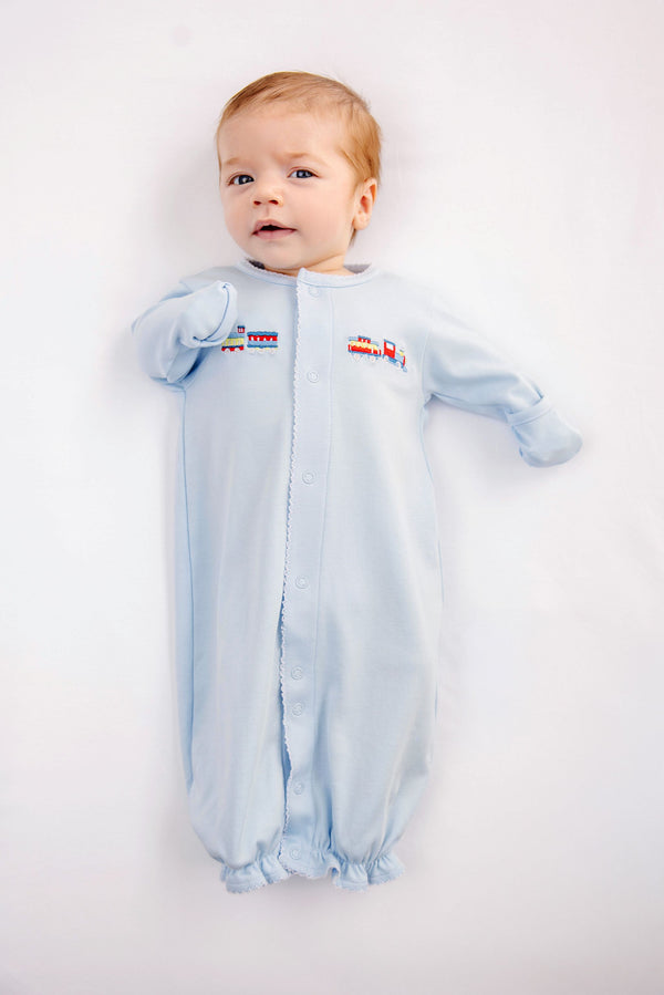 A baby wearing The Beaufort Bonnet Company’s Twinkle Twinkle 2-in-1 - Buckhead Blue with Trains outfit lies on a white surface, looking up with a slight smile and one arm raised.