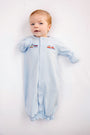 A baby wearing The Beaufort Bonnet Company’s Twinkle Twinkle 2-in-1 - Buckhead Blue with Trains outfit lies on a white surface, looking up with a slight smile and one arm raised.