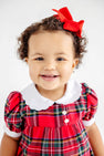 A smiling toddler wears a Mary Baker Bubble - Society Prep Plaid with Worth Avenue White, featuring a red plaid bubble romper, white scalloped collar, and matching red hair bow for a classic holiday baby outfit.