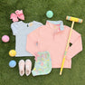 Children’s outfit laid out on grass features The Beaufort Bonnet Company’s Prepletic™ Hayword Half-Zip in Palm Beach Pink with Palm Beach Pink Stork, a light blue shirt, floral shorts, pink shoes, a pink bow, pastel balls, and a wooden croquet mallet.