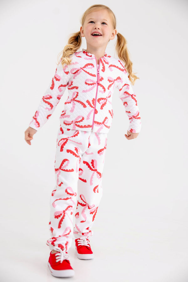 A young girl wears a white, quilted Georganna Jacket - Quilted Bonnie Scot Bows and matching joggers patterned with pink and red bows, paired with red sneakers.