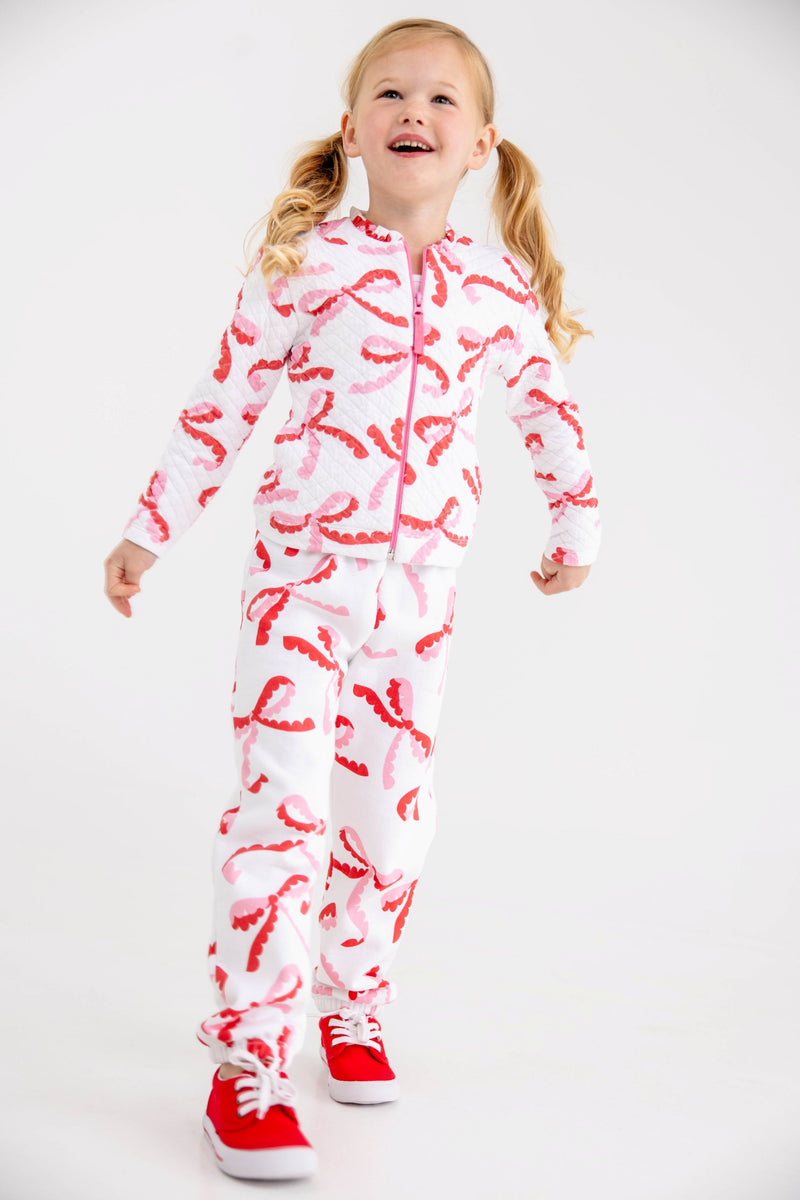 A young girl wears a white, quilted Georganna Jacket - Quilted Bonnie Scot Bows and matching joggers patterned with pink and red bows, paired with red sneakers.