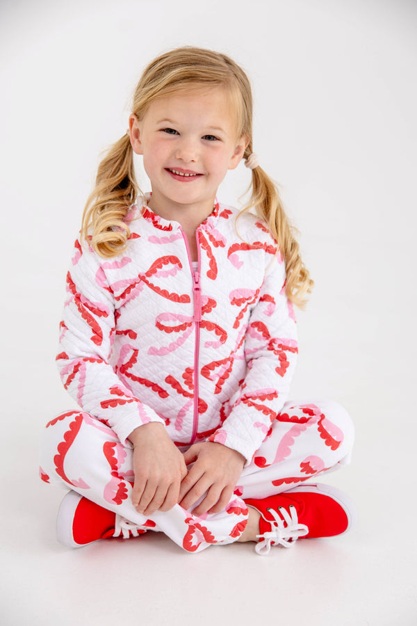 A young girl sits cross-legged wearing a white, quilted Georganna Jacket - Quilted Bonnie Scot Bows and matching pants featuring a pink and red bow pattern, paired with red sneakers.