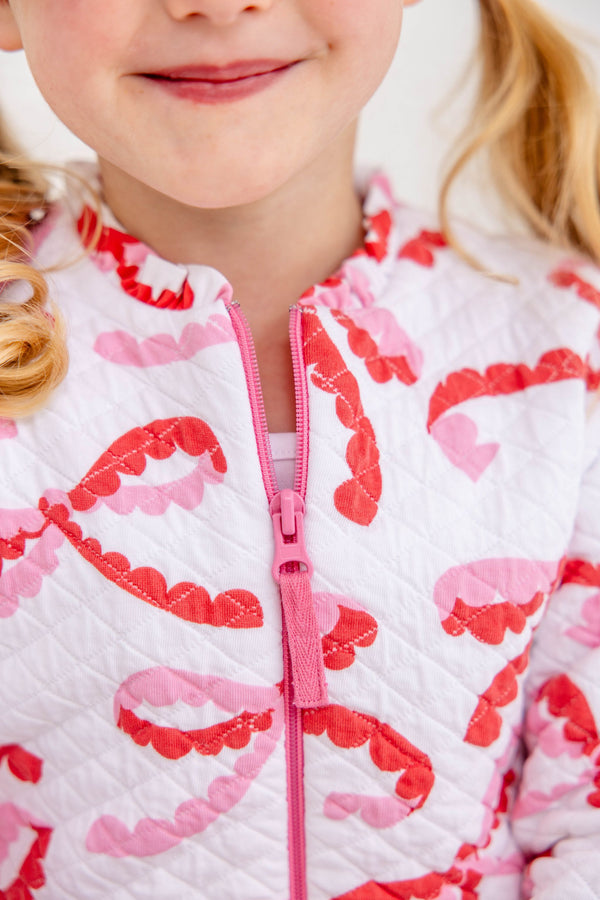 A child wears a Georganna Jacket - Quilted Bonnie Scot Bows, a white quilted piece featuring a repeating pattern of pink and red bow shapes with a pink front zipper.