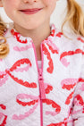 A child wears a Georganna Jacket - Quilted Bonnie Scot Bows, a white quilted piece featuring a repeating pattern of pink and red bow shapes with a pink front zipper.