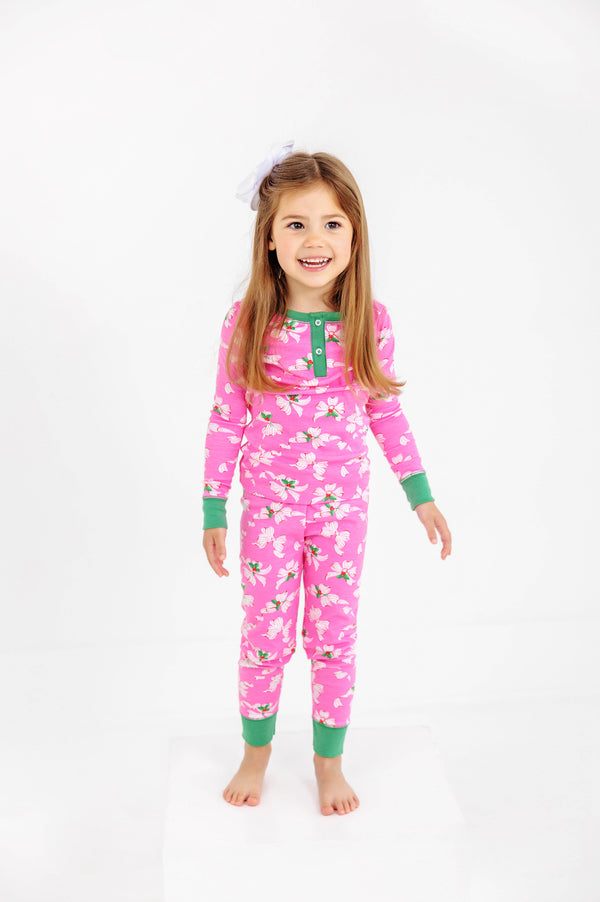 A young girl with long hair is smiling against a plain white background, wearing The Beaufort Bonnet Company’s Sara Jane’s Sweet Dream Set - Bows of Holly with Kiawah Kelly Green, a pink holiday family matching pajama set.