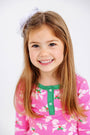 A young girl with long brown hair, wearing The Beaufort Bonnet Company’s Sara Jane’s Sweet Dream Set in the Bows of Holly with Kiawah Kelly Green print, smiles at the camera. She has a large white bow in her hair and stands against a plain white background, looking cozy and comfortable in her favorite Pima cotton pajamas.