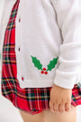 A child is shown wearing The Beaufort Bonnet Company’s Cambridge Cardigan - Worth Avenue White with Holly, featuring embroidered holly leaves and red berries, layered over a red plaid dress. Only the lower half of the child’s face and body is visible.
