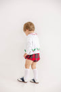 A young child stands facing a white wall, wearing The Beaufort Bonnet Company’s Cambridge Cardigan in Worth Avenue White with Holly.