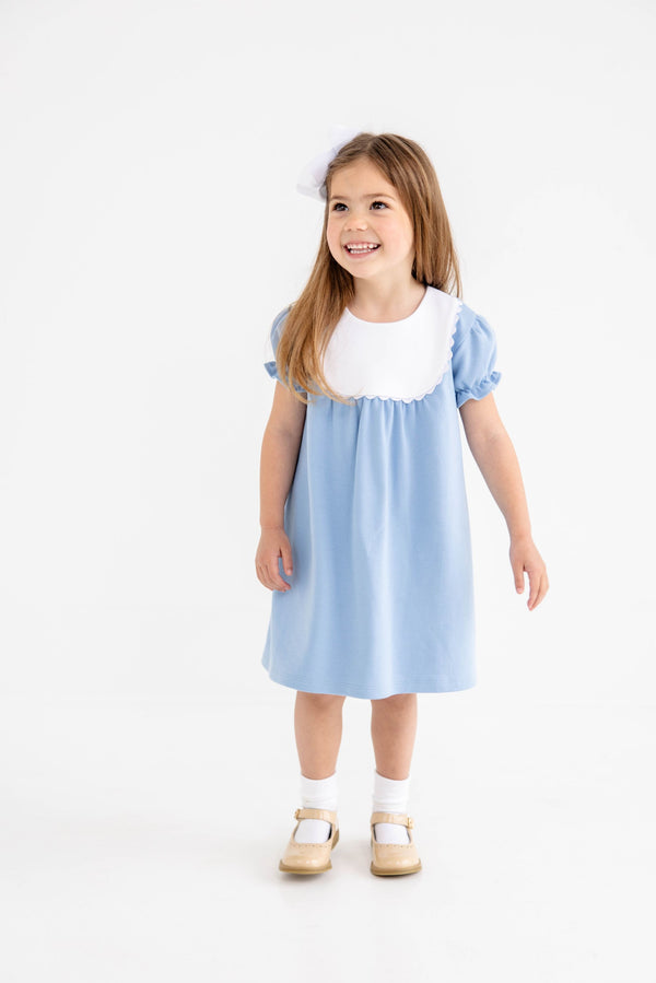 A young girl with long brown hair wears The Beaufort Bonnet Company’s Pharish Frock in Barrington Blue with Worth Avenue White, paired with white socks, tan shoes, and a white bow in her hair. She is smiling and standing against a plain white background.