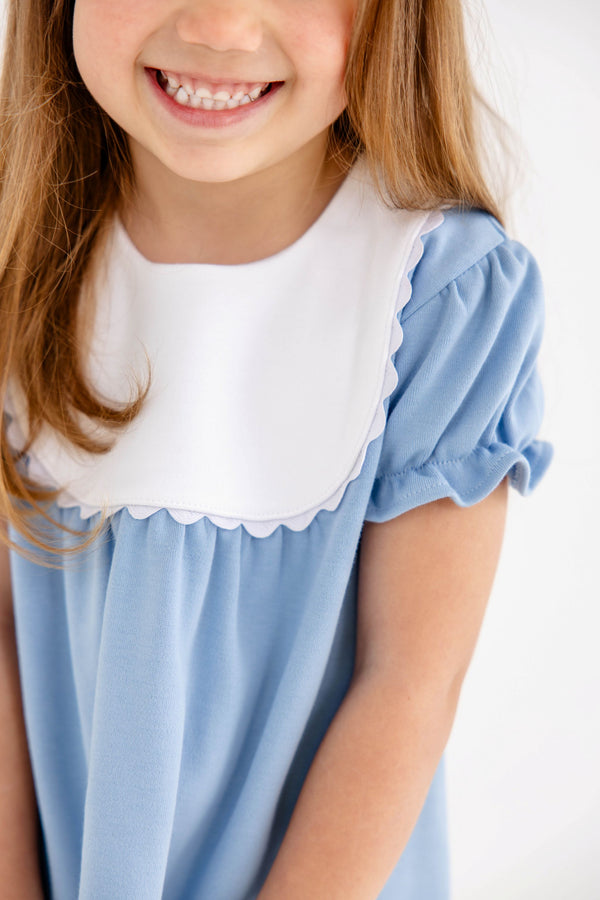 A young girl smiles while wearing The Beaufort Bonnet Company’s Pharish Frock in Barrington Blue with Worth Avenue White. The dress features puff sleeves and a white scalloped collar. Her face is partially out of frame, and her long brown hair falls loosely over her shoulders.