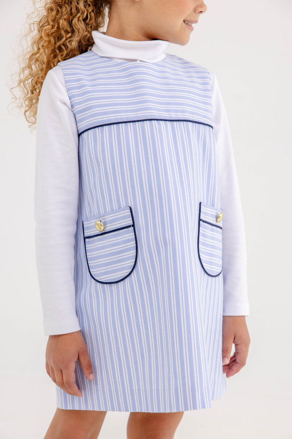 A child wears a white long-sleeve shirt under a Jill Jumper - Park City Periwinkle Stripe with Nantucket Navy, a light blue and white striped dress featuring two front pockets with navy trim and gold buttons.