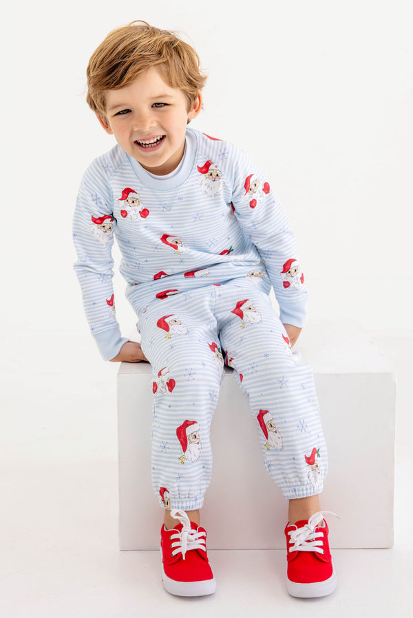 A smiling young boy wearing light blue and white striped Gates Sweeney Sweatpants - Dear Santa (Buckhead Blue Stripe) with a Santa Claus print and red sneakers.
