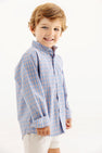 A smiling young boy with light brown hair wears The Beaufort Bonnet Company’s Dean’s List Dress Shirt in Wheaton Windowpane with Barrington Blue Stork, paired with white shorts, standing against a plain white background.