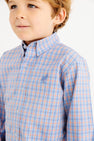 A young child wearing The Beaufort Bonnet Company’s Dean’s List Dress Shirt in Wheaton Windowpane with Barrington Blue Stork, featuring a light blue and orange windowpane pattern and a small embroidered stork logo on the chest, stands against a white background.