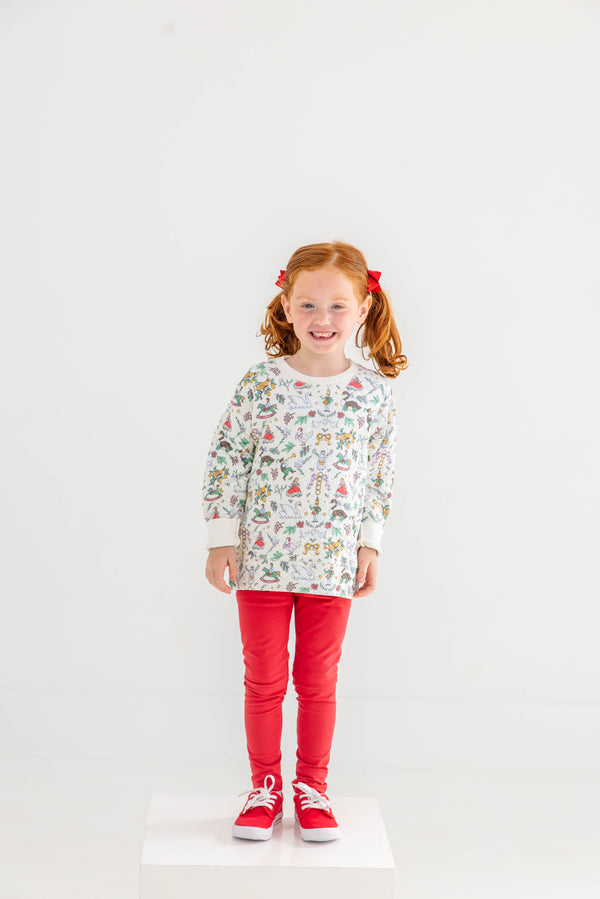 A smiling young girl with red hair in pigtails stands on a white platform, wearing The Beaufort Bonnet Company’s Cassidy Comfy Crewneck - 12 Days of Christmas, paired with red pants, red bows in her hair, and red sneakers, against a plain white background.