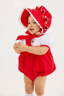 A young child stands against a plain light background, smiling and holding something in their hands, dressed in The Beaufort Bonnet Company’s Cindy Lou Bubble in Richmond Red & Worth Avenue White with Christmas Tree embroidery and a coordinating red bonnet.