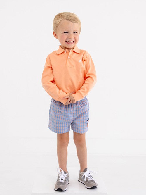 A smiling young boy with short blond hair is wearing The Beaufort Bonnet Company’s Sheffield Shorts in Wheaton Windowpane with a Seashore Sherbet Stork & Pumpkin Applique, paired with an orange long-sleeve polo shirt and gray sneakers, standing against a plain white background.