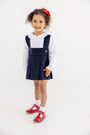 A young girl wears a Pearl Prep Skirt - Nantucket Navy Corduroy with Society Prep Plaid over a long-sleeved white collared shirt, paired with white socks and red patent leather T-strap shoes in a classic Society Prep style.