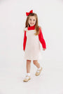 A smiling young girl with long hair stands against a plain white background, wearing a red shirt and The Beaufort Bonnet Company’s Julia Jumper in Palmetto Pearl & Richmond Red Microdot Corduroy, styled with white socks, tan shoes, and a large red bow in her hair.