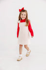 A smiling young girl with long hair stands against a plain white background, wearing a red shirt and The Beaufort Bonnet Company’s Julia Jumper in Palmetto Pearl & Richmond Red Microdot Corduroy, styled with white socks, tan shoes, and a large red bow in her hair.