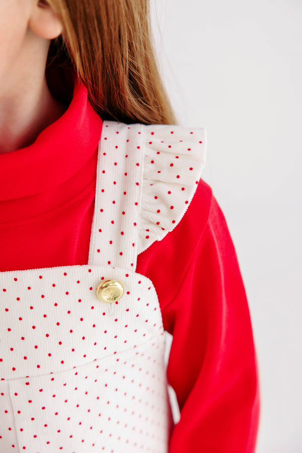 Close-up of a child wearing a vibrant red turtleneck and The Beaufort Bonnet Company’s Julia Jumper—crafted in Palmetto Pearl & Richmond Red Microdot corduroy, featuring gold buttons and ruffled shoulder detailing. Only part of the child’s face and hair is shown, perfectly capturing the festive spirit of this holiday girls’ dress.