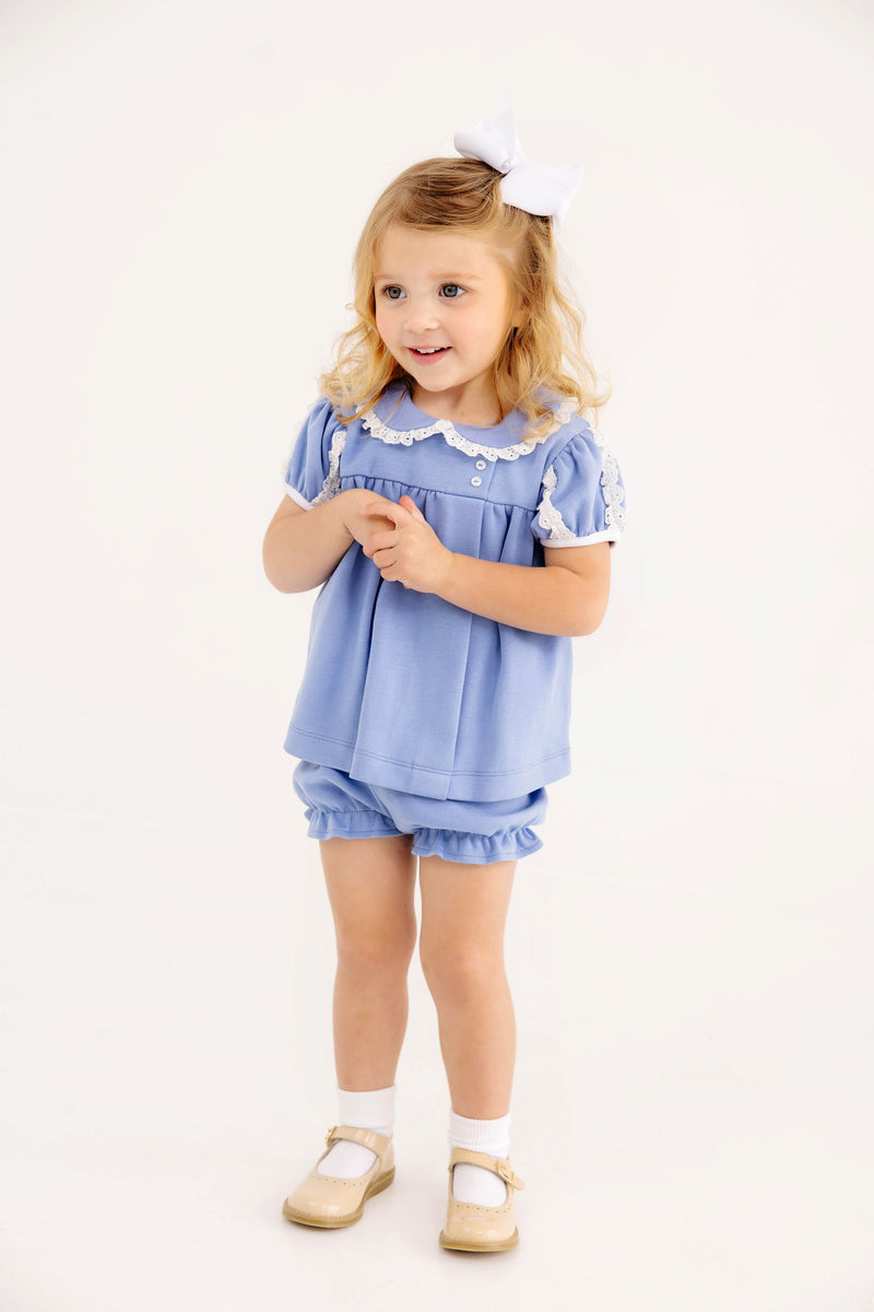 A young girl wears a Mary Baker Bloomer Set - Park City Periwinkle with Worth Avenue White, featuring a white lace-trimmed collar, ruffled sleeves, white socks, and cream-colored shoes.
