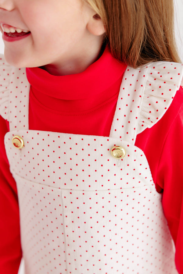 A child wearing The Beaufort Bonnet Company’s Mitzy Sue Slacks in Richmond Red and a white pinafore dress with red polka dots and ruffled straps, smiling with only the lower half of the face visible.