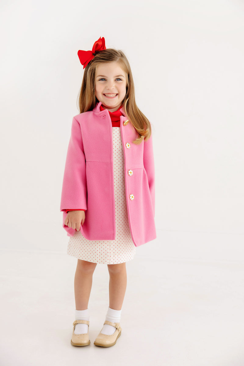 A young girl smiles while wearing The Beaufort Bonnet Company’s Penelope Peacoat in Hamptons Hot Pink with Richmond Red, paired with a white dress featuring small dots, white socks, tan shoes, and a red bow in her hair. The background is plain and white.