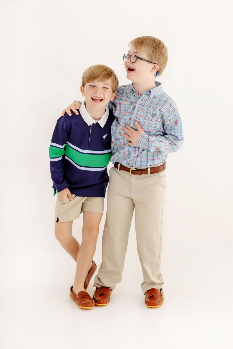 Two smiling boys stand together. One wears a navy sweater with green and white stripes paired with shorts, while the other wears glasses and The Beaufort Bonnet Company’s Dean’s List Dress Shirt in Kensington Court Plaid with Barbados Blue Stork, along with khaki pants. Both sport brown loafers. They appear happy and close.