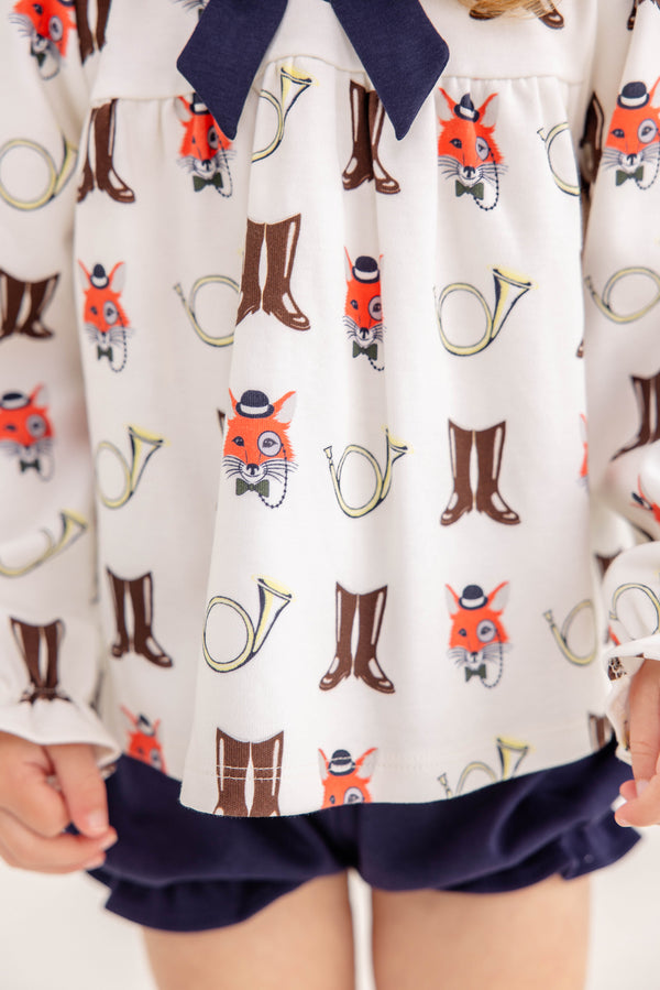 A child wears The Beaufort Bonnet Company’s Long Sleeve Banks Bow Bloomer Set in the Happy Hunting with Palmetto Pearl & Nantucket Navy print. This fall ensemble—featuring a top with whimsical prints of fox faces, brown boots, and yellow French horns paired with navy blue bloomers—is perfect as baby girl autumn clothing. Only the lower torso and hands are visible.