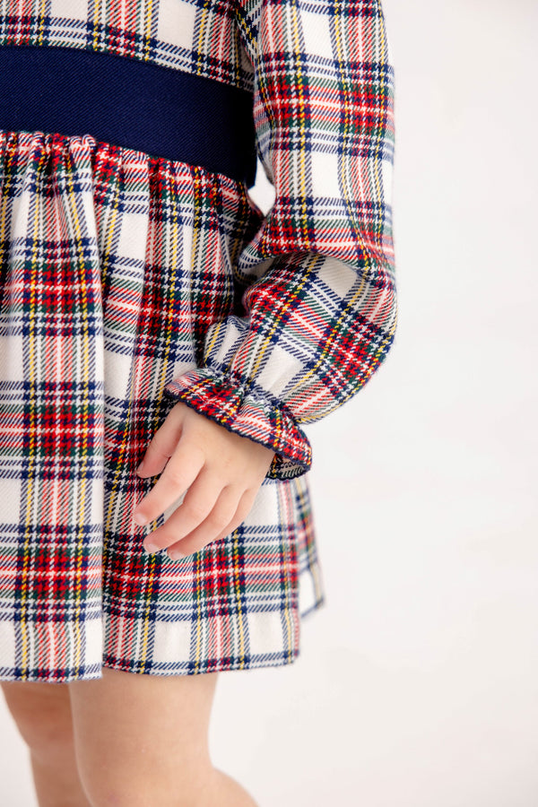 A child wears a Long Sleeve Cindy Lou Sash Dress - Paris Pike Plaid with Nantucket Navy, featuring a navy blue sash at the waist and ruffled sleeve cuffs, perfect as a holiday party dress.