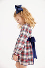 A young girl wearing a Long Sleeve Cindy Lou Sash Dress - Paris Pike Plaid with Nantucket Navy, featuring a navy blue sash and matching hair bow, perfect for a holiday party.