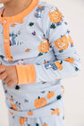 A child wearing The Beaufort Bonnet Company’s Payton Pajama Set - Candy & Critters (Buckhead Blue) with Seashore Sherbet, featuring a playful pattern of smiling pumpkins, raccoons, and autumn leaves.