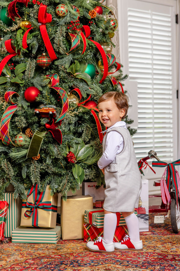 A young boy wearing a Jack Keene Jon Jon - Grantley Gray Houndstooth with Grantley Gray, a white shirt, and red and white shoes stands smiling in front of a decorated Christmas tree for a winter family portrait.