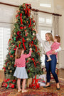 A woman and two young children decorate a Christmas tree with ornaments and ribbons as wrapped gifts sit beneath the branches. One child wears the Reece Ruffle Sweater in Pier Party Pink & Richmond Red Stripe by The Beaufort Bonnet Company, while the other sports a pink striped sweater. The joyful scene takes place in a bright, festively decorated room.