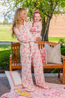 A woman and young girl, both in matching floral pajamas, smile on a wooden bench outside. The girl has a pink bow in her hair. Decorative pillows and The Beaufort Bonnet Company’s Silent Night Throw - Gourd-geous (Palmetto Pearl) with Romany Rose cozy blanket are on the ground in front of them, along with some toys.