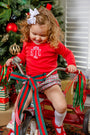 A young child with curly hair and a white bow is dressed in The Beaufort Bonnet Company’s Natalie Knickers in Wicker Park Plaid with Richmond Red, riding a tricycle adorned with red and green ribbons in front of a Christmas tree decorated with ornaments and wrapped gifts.