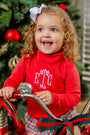 A smiling young girl with curly hair and a white bow sits on a tricycle in front of a decorated Christmas tree, wearing The Beaufort Bonnet Company’s Mitzy Sue Slacks in Richmond Red with white monogram letters and plaid accents.