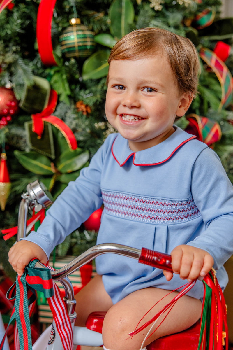 A smiling toddler wears a Long Sleeve Bradford Bubble - Barrington Blue with Richmond Red Smocking while sitting on a red tricycle in front of a Christmas tree.