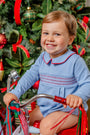 A smiling toddler wears a Long Sleeve Bradford Bubble - Barrington Blue with Richmond Red Smocking while sitting on a red tricycle in front of a Christmas tree.
