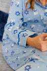 A person wearing The Beaufort Bonnet Company’s “Let Me Lounge Set - Eight Nights to Remember with Barbados Blue,” featuring blue Stars of David, dreidels, and menorahs, is sitting on a couch, showing only their torso and hand.