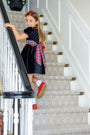A young girl in the Cindy Lou Sash Dress – Newport Night Velveteen with Bucktown Tartan by The Beaufort Bonnet Company smiles as she walks up a carpeted staircase, holding the railing.
