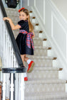 A young girl in the Cindy Lou Sash Dress – Newport Night Velveteen with Bucktown Tartan by The Beaufort Bonnet Company smiles as she walks up a carpeted staircase, holding the railing.