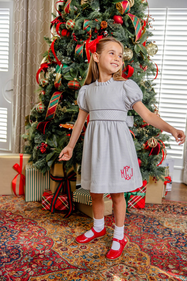 A young girl stands in front of a Christmas tree wearing a Cindy Lou Sash Dress - Grantley Gray Houndstooth with short puff sleeves, a red monogram on the skirt, and red Mary Jane shoes.