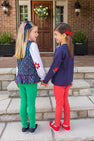 Two young girls holding hands and smiling at each other, wearing Kiawah Kelly Green sweaters and Mitzy Sue Slacks while standing on a brick house porch.