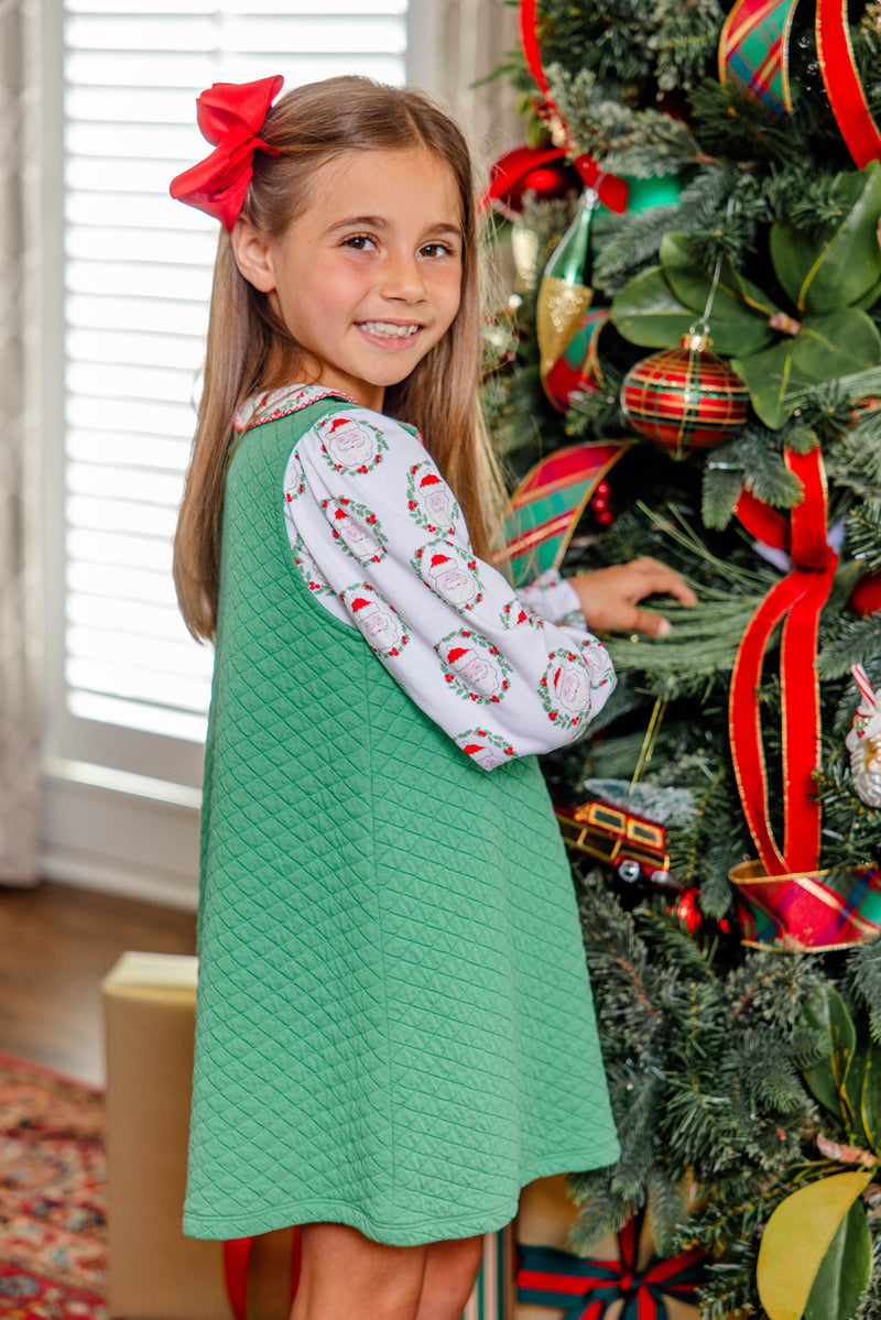 A smiling young girl stands by a decorated Christmas tree adorned with red ribbons and ornaments, dressed for the holidays in The Beaufort Bonnet Company’s Long Sleeve Maude’s Peter Pan Collar Shirt – North Pole Portrait with Richmond Red.